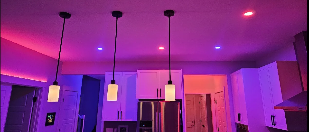A kitchen at night in a pink and violet scene: pendants, pot lights, cove lighting and a wall panel by the door