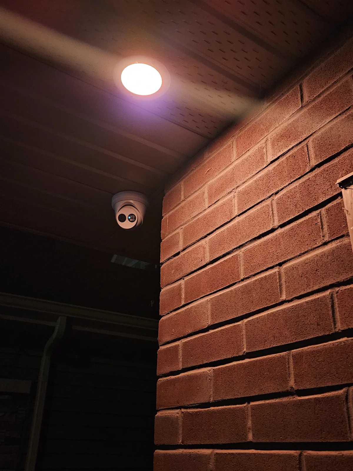 A dome camera and warm pot light in the soffit beside a brick wall at night